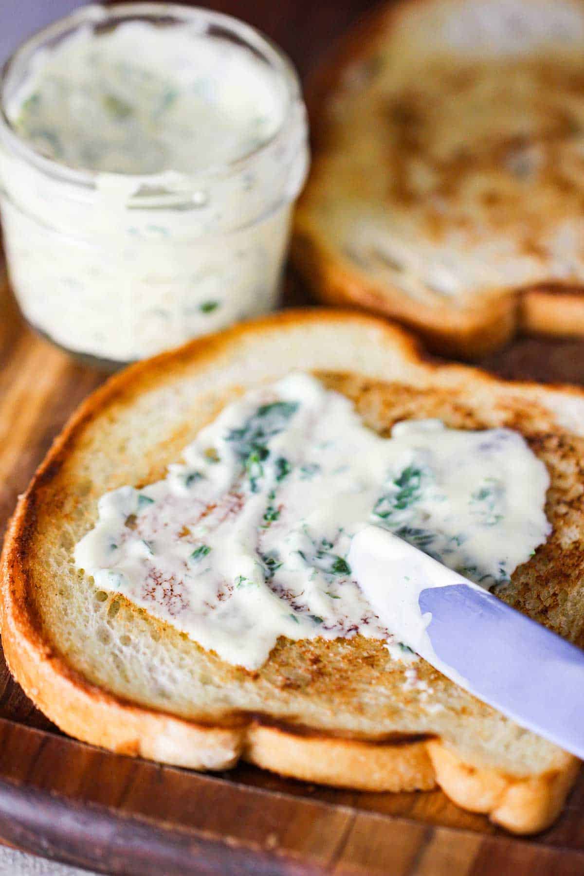 A person smearing garlic basil aioli over a sliced of toasted bread next to a small bowl filled with aioli.