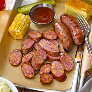 A close-up view of a metal trey lined with brown butcher paper filled with a serving of sliced Homemade Jalapeño Cheddar Sausage with a piece of corn on the cob, BBQ sauce, a pickle spear, and a square of corn bread all surrounding the sausage.