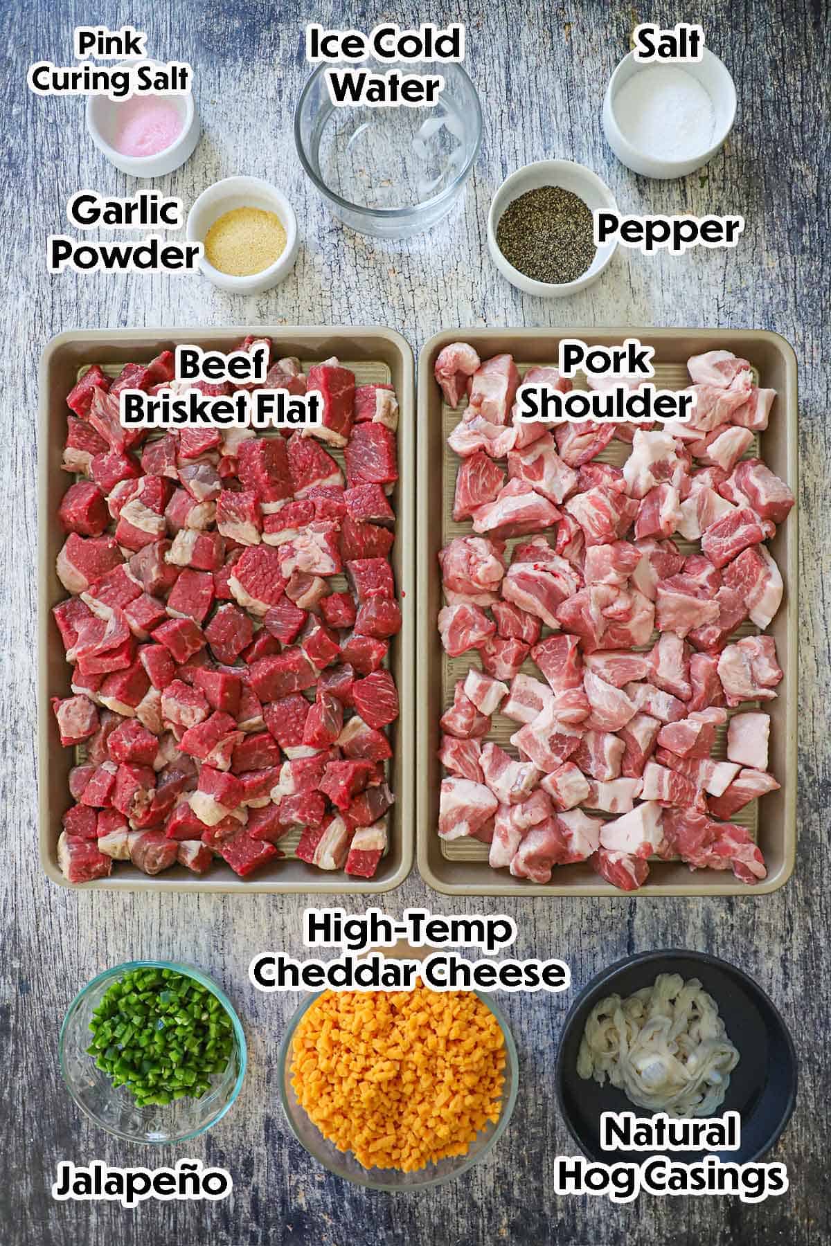 An arrangement of ingredients for Homemade Texas Jalapeño Cheddar Sausage on a grey wooden background included cubed beef brisket, pork shoulder, high-temperature cheddar cheese cubes, jalapeño, hog casings, curing salt, and seasonings.