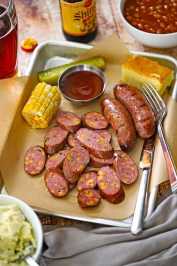 A metal trey lined with brown butcher paper filled with a serving of sliced Homemade Jalapeño Cheddar Sausage with a piece of corn on the cob, BBQ sauce, a pickle spear, and a square of corn bread all surrounding the sausage.