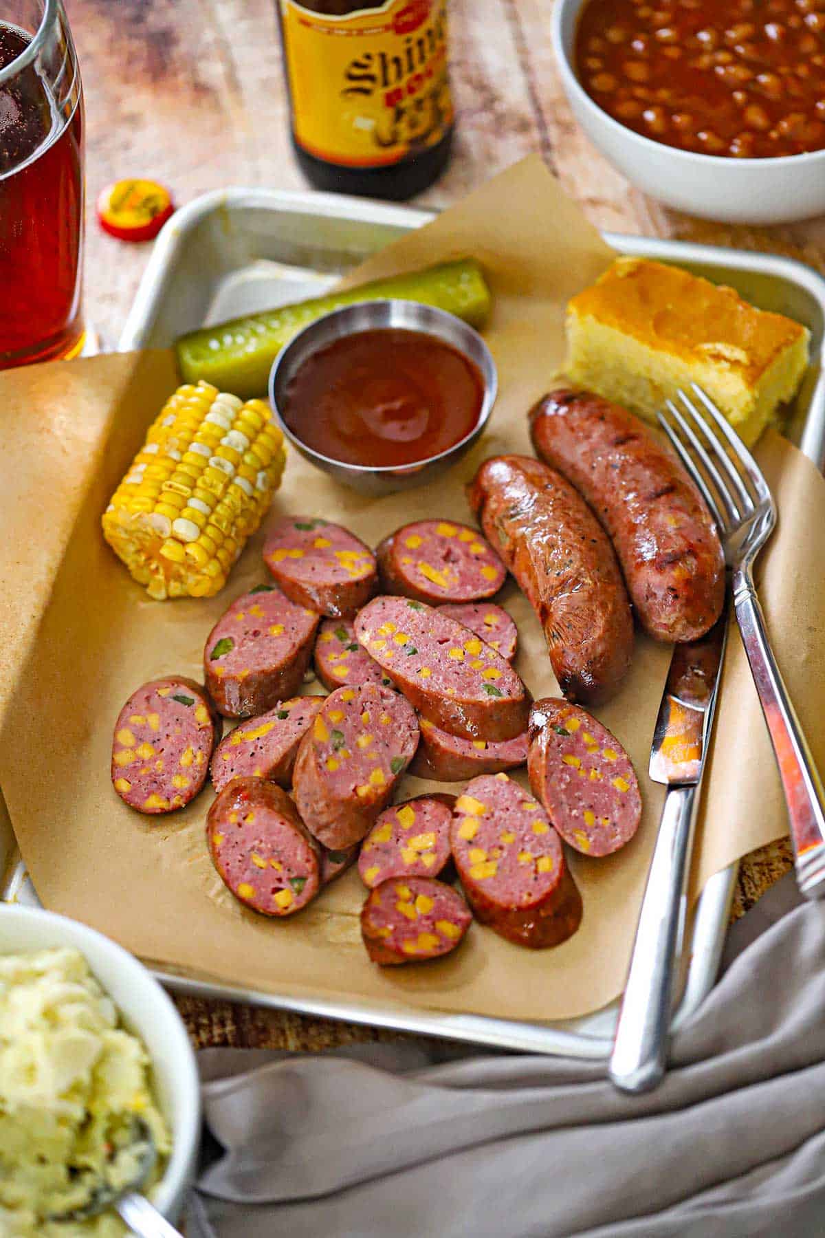 A metal trey lined with brown butcher paper filled with a serving of sliced Homemade Jalape&ntilde;o Cheddar Sausage with a piece of corn on the cob, BBQ sauce, a pickle spear, and a square of corn bread all surrounding the sausage.