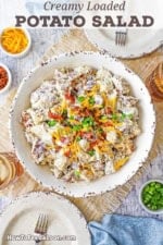 An overhead view of a white serving bowl filled with a creamy loaded potato salad topped with crispy bacon bits, chopped scallions, and shredded cheddar cheese and surrounded by small bowls filled with cheese, bacon, and scallions.