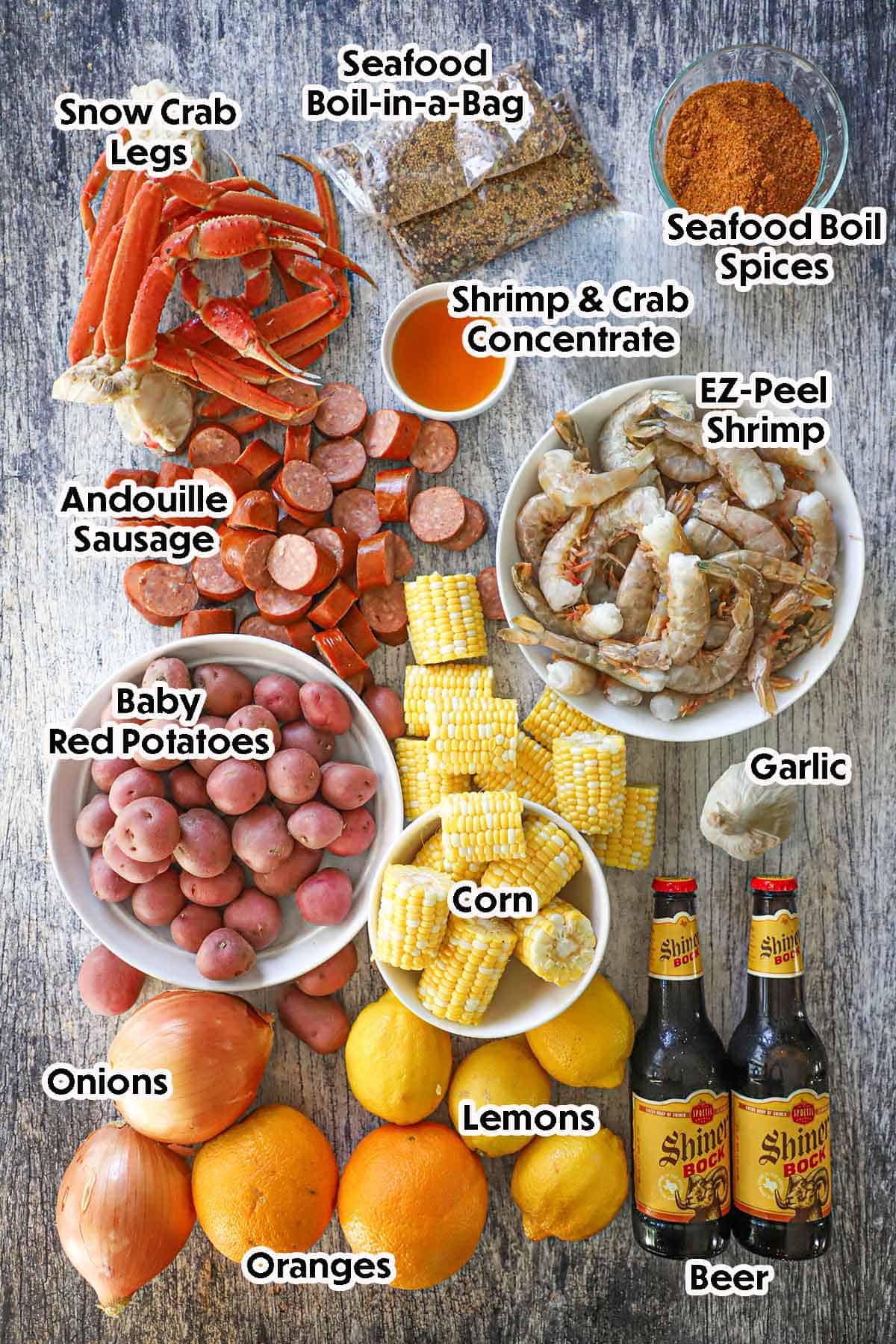 An arrangement of ingredients for Louisiana Seafood Boil on a grey wooden background including EZ-peel shrimp, corn on the cob, red potatoes, crab legs, onions, lemons, oranges, garlic, beer, and seasonings.
