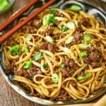 A close-up view of a shallow bowl filled with a serving of Mongolian Ground Beef Noodles garnished with chopped scallions and a pair of chop sticks resting on the side of the bowl.