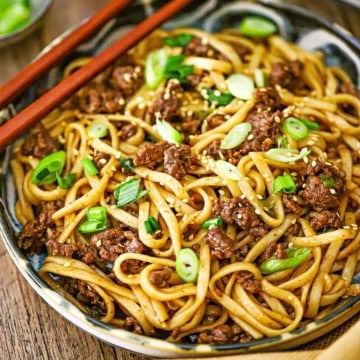 A close-up view of a shallow bowl filled with a serving of Mongolian Ground Beef Noodles garnished with chopped scallions and a pair of chop sticks resting on the side of the bowl.
