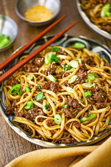 A straight-on view of a shallow bowl filled with a serving of Mongolian Ground Beef Noodles garnished with chopped scallions and a pair of chop sticks resting on the side of the bowl.