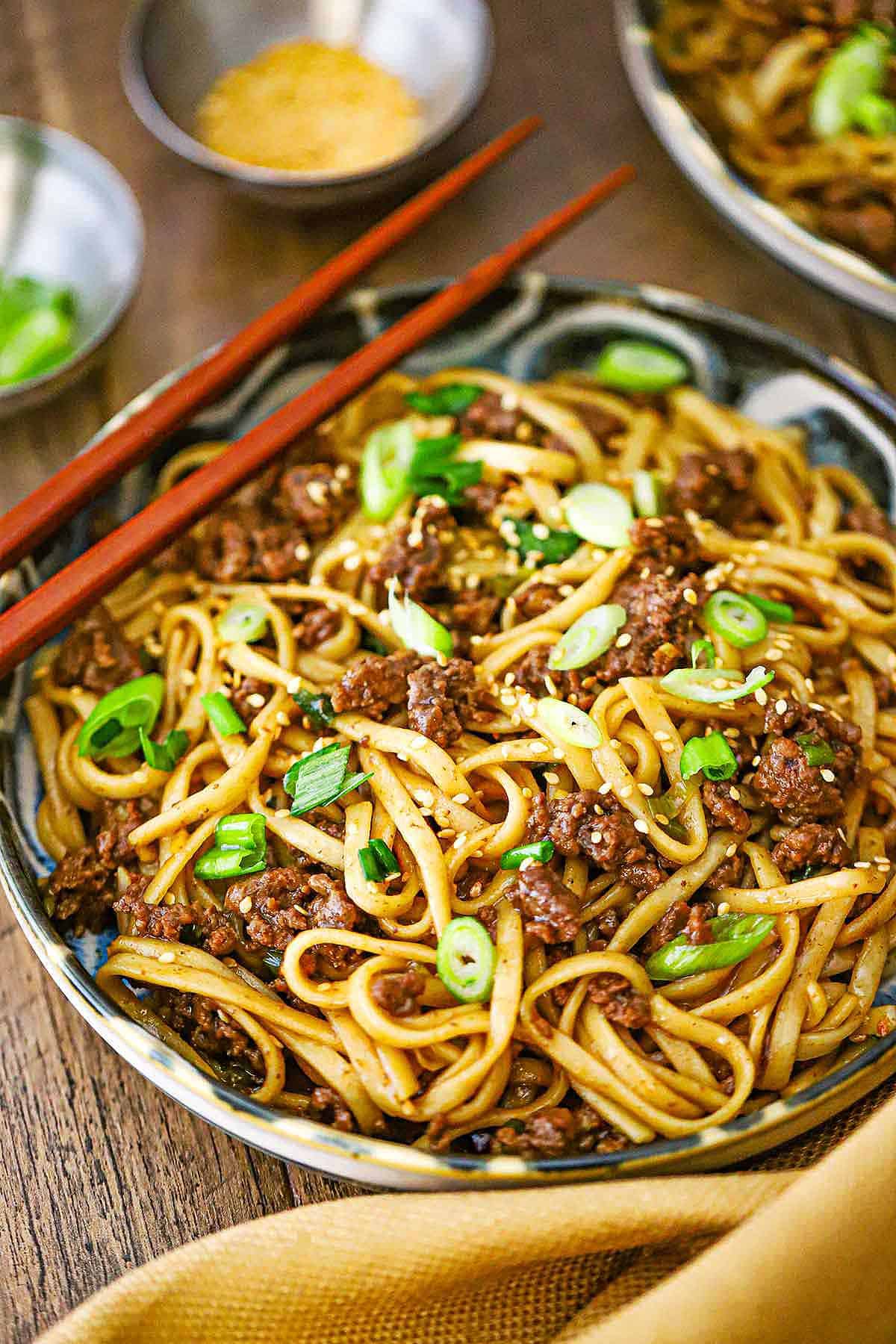 A straight-on view of a shallow bowl filled with a serving of Mongolian Ground Beef Noodles garnished with chopped scallions and a pair of chop sticks resting on the side of the bowl.