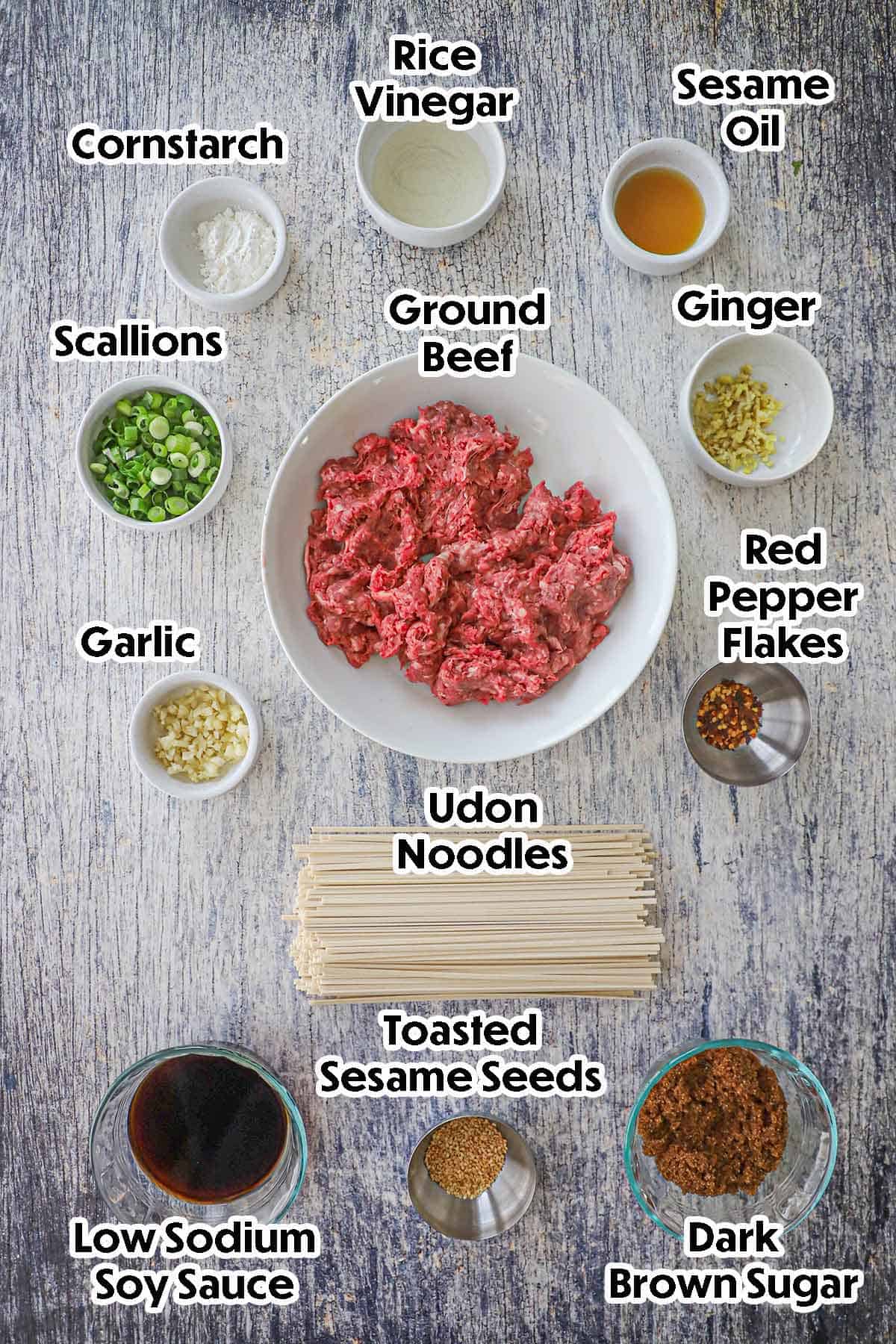 An arrangement of ingredients for Mongolian Ground Beef Noodles on a wooden grey background including uncooked ground beef, brown sugar, soy sauce, garlic, scallions, minced ginger, oil, vinegar, cornstarch, and seasonings.