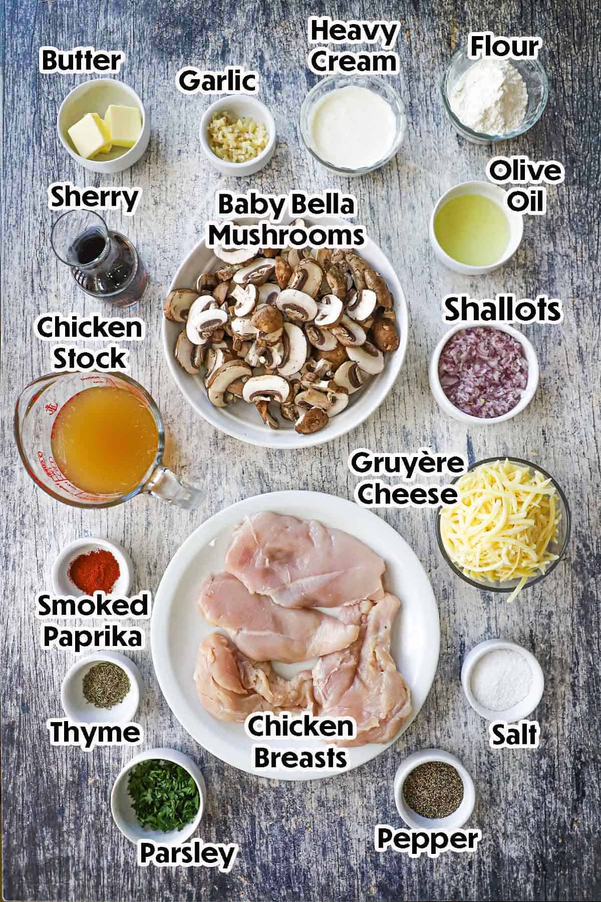An arrangement of ingredients for chicken Gloria recipe on a grey wooden background including raw chicken breast, sliced mushrooms, shallots, Sherry wine, garlic, flour, butter, shredded cheese, chicken stock, oil, herbs and seasonings.