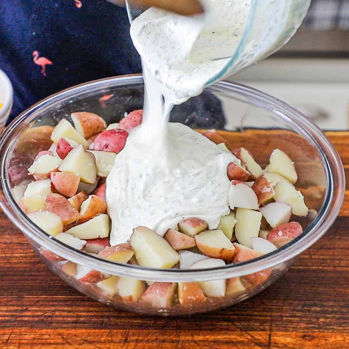 A person pouring a creamy potato salad dressing over the top of tender pieces of red potatoes in a large glass bowl.