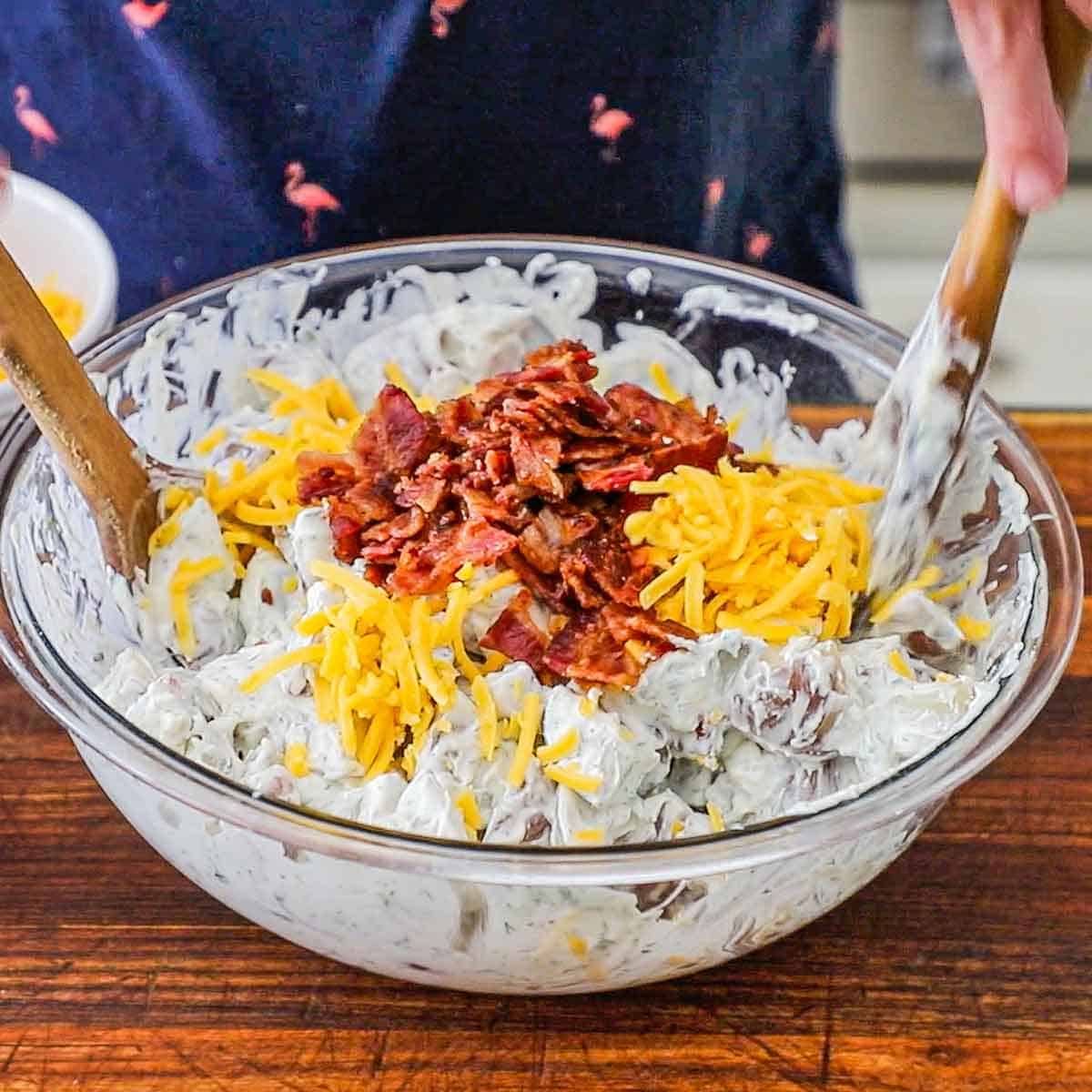 A person using two large wooden spoons to fold crispy bacon bits and shredded cheddar cheese into chunks of tender potatoes that are coating with a creamy herb dressing.