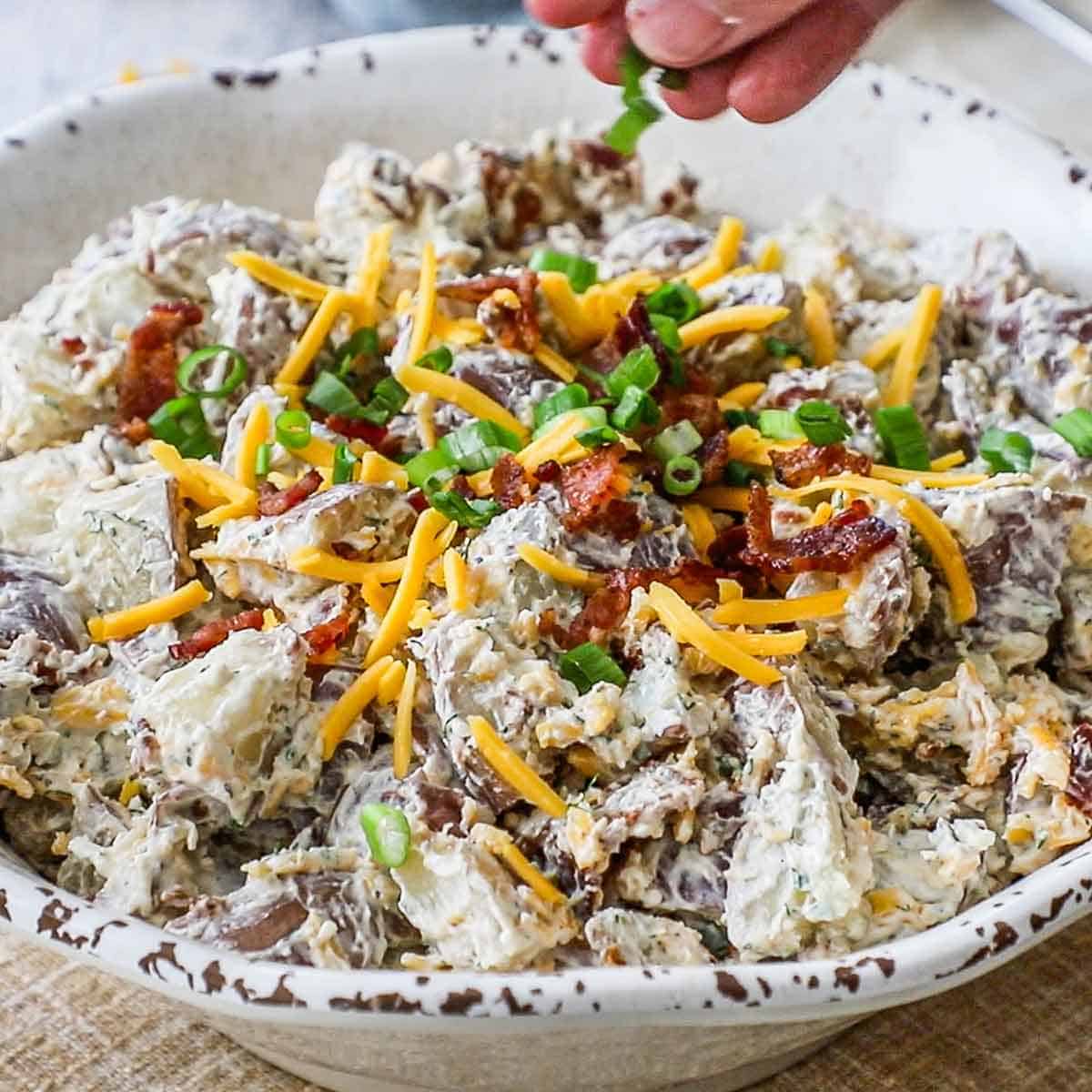 A person sprinkling chopped chives over the top of a creamy loaded potato salad in a large white bowl.