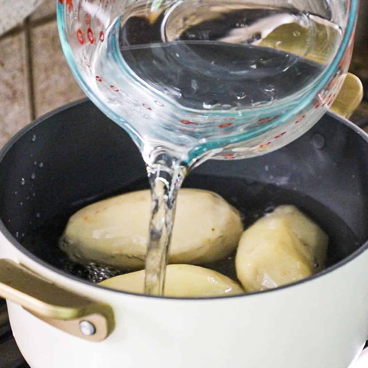 A person pouring cool water from a large glass measuring cup into a pot on a gas stove that is filled with three large peeled russet potatoes.