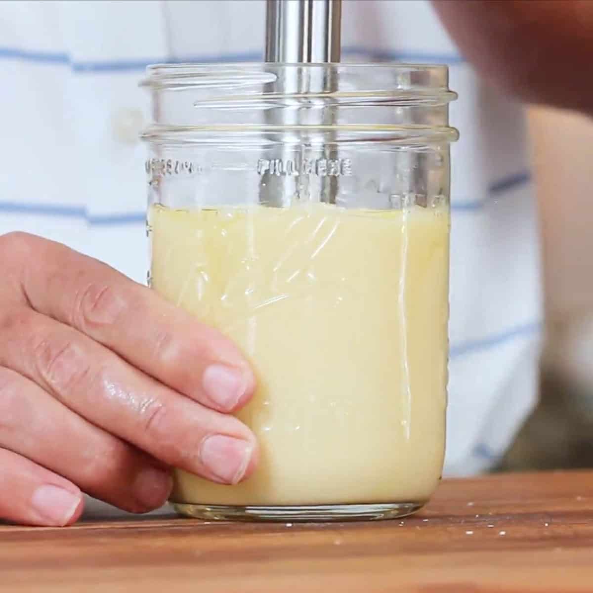 A person using an immersion blender to create homemade mayonnaise in a Mason jar on a wooden cutting board.