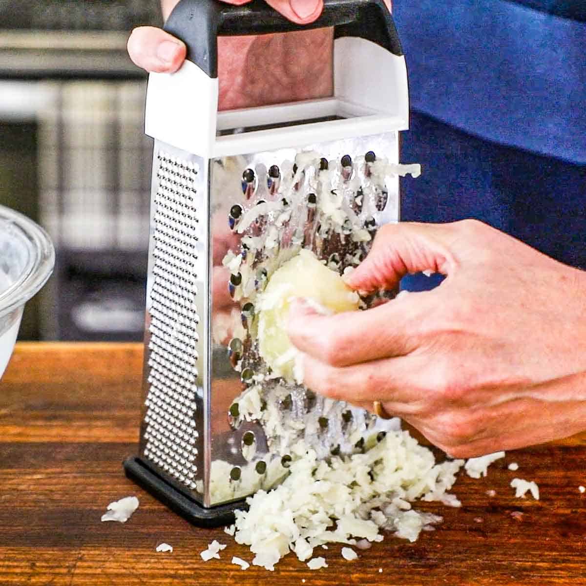 A person grating a par-boiled peeled russet potato on a box grater on a wooden cutting board.