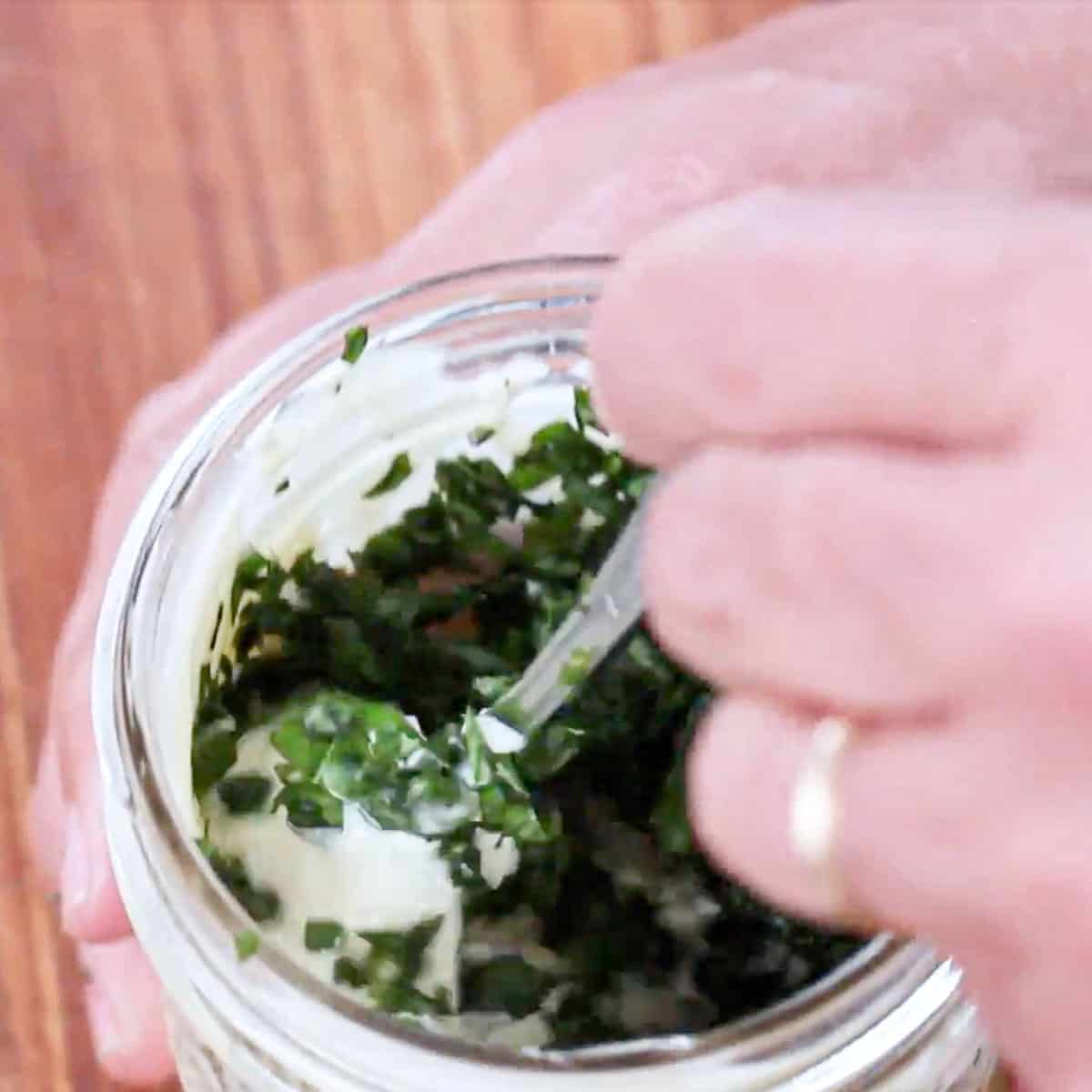 A person using a spoon to fold chopped fresh basil and minced garlic into a jar filled with homemade mayonnaise.