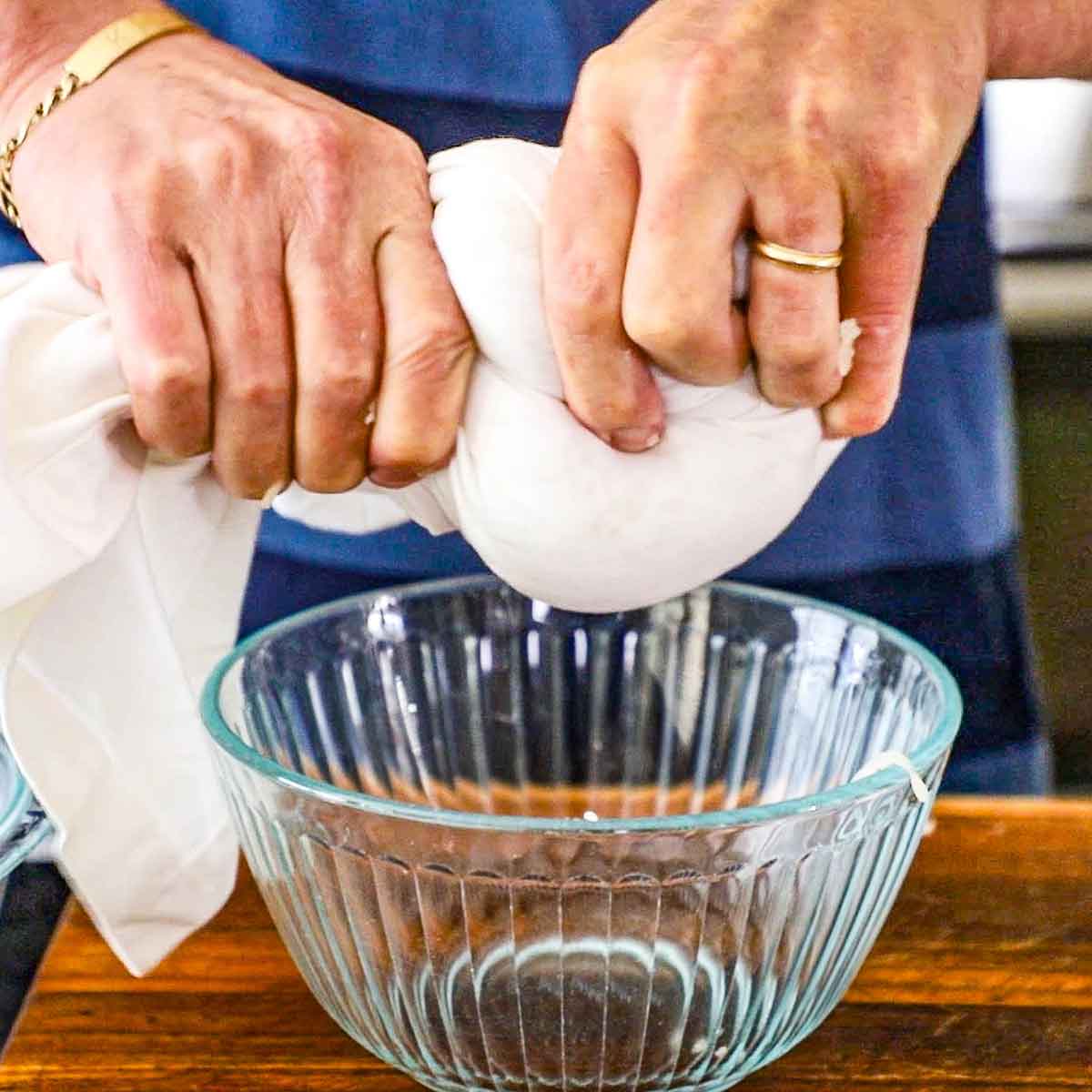 A person wringing a white cloth that if filled with grated par-boiled potatoes over a glass bowl.