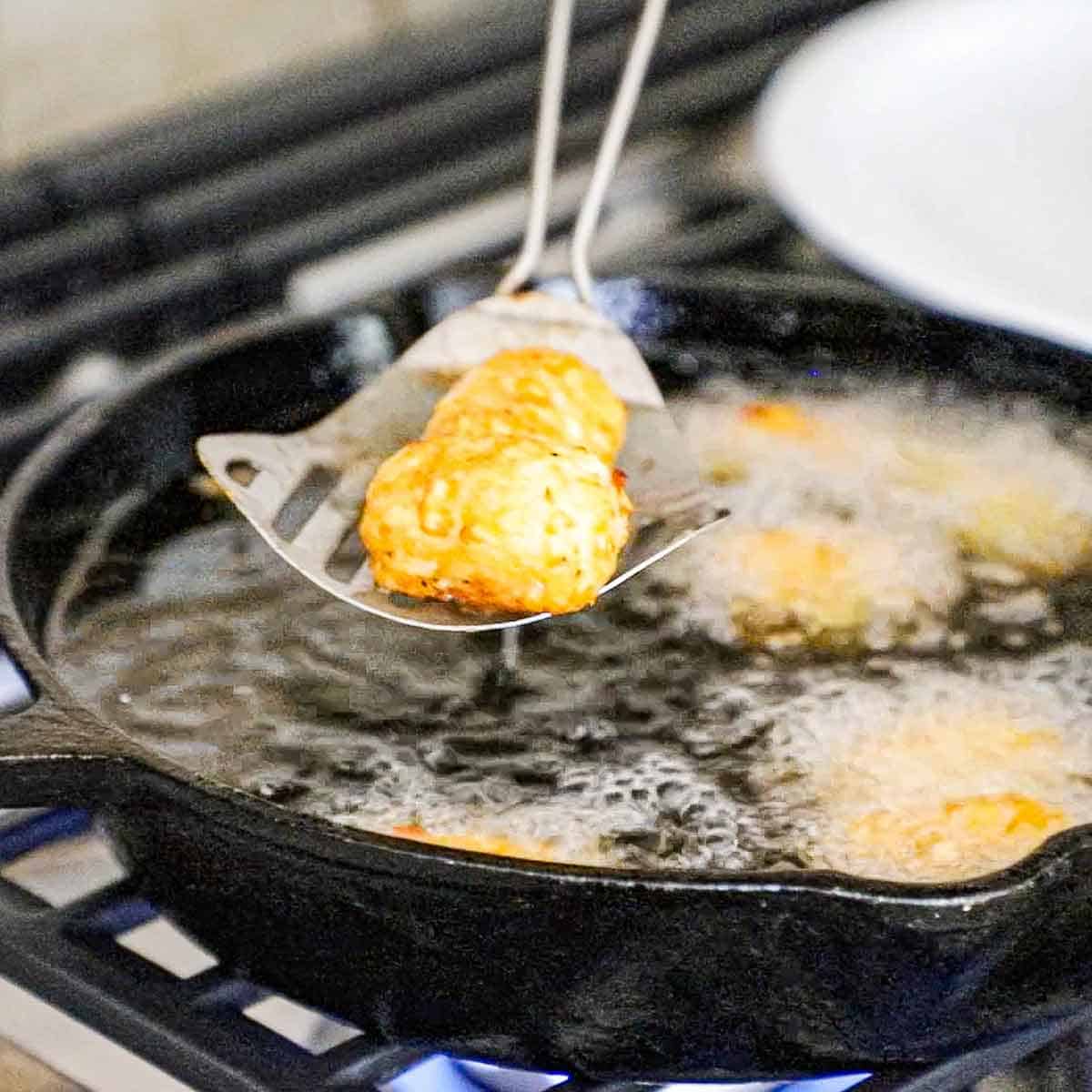 A person using a metal slotted spatula to raise two fried homemade tater tots out of a cast-iron skillet that is filled with hot oil and other tater tots frying in the oil.