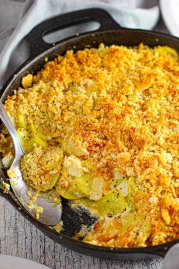 A close-up view of a large cast-iron skillet filled with a serving of summer squash casserole with a portion of it missing with a serving spoon in the pan.