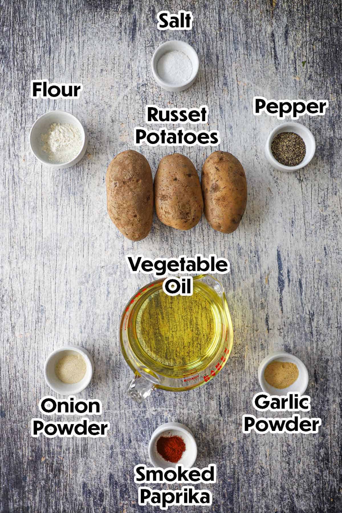 An arrangement of ingredients for homemade tater tots on a grey wooden background including Russet potatoes, vegetable oil, flour, and seasonings.