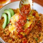 A person using a spoon to drizzle a baked salsa sauce over the top of Easy Baked Salsa Chicken resting on a bed of Mexican rice in a shallow bowl.