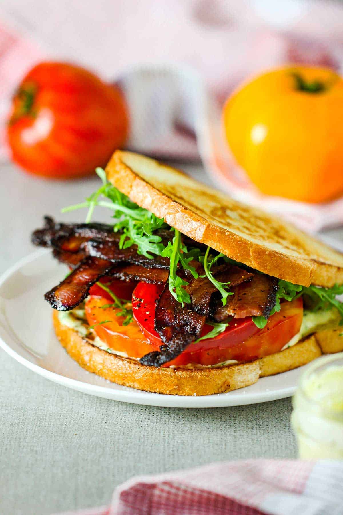 A straight-on view of a Classic BLT with Garlic Basil Aioli on lightly toasted sourdough bread on a white plate with a red and yellow tomato in background.