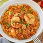 An overhead view of a white bowl filled with a serving of authentic jambalaya topped with seared shrimp, andouille sausage, and chopped scallions.