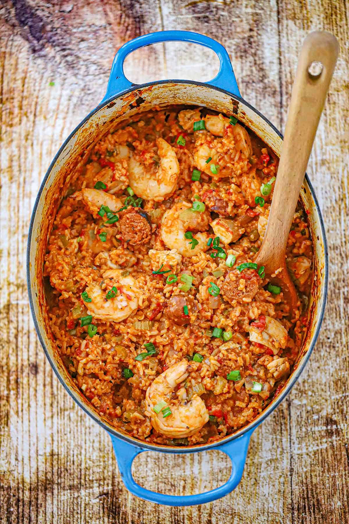 An overhead view of a large oval Dutch oven filled with a freshly prepare authentic jambalaya with a wooden spoon inserted into the dish.