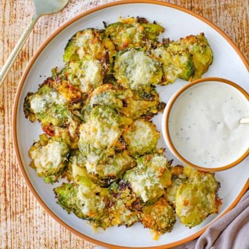 An close-up overhead view of a round white plate filled with a serving of Parmesan Smashed Brussels Sprouts in a pile next to a round bowl filled with homemade Ranch dressing.