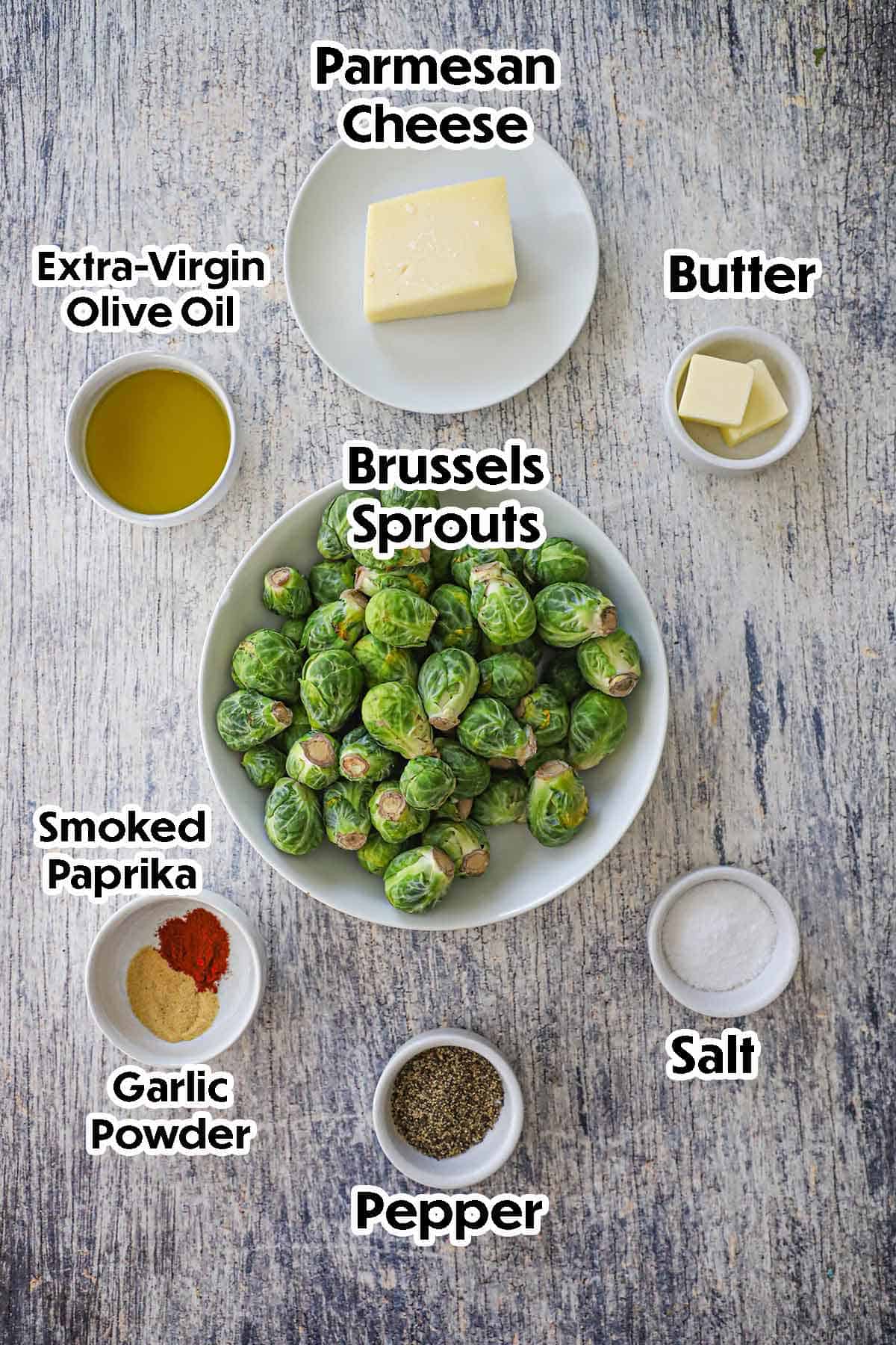 An arrangement of ingredients for Parmesan Smashed Brussels Sprouts on a grey wooden background including Brussels sprouts, olive oil, butter, Parmesan cheese, and seasonings.