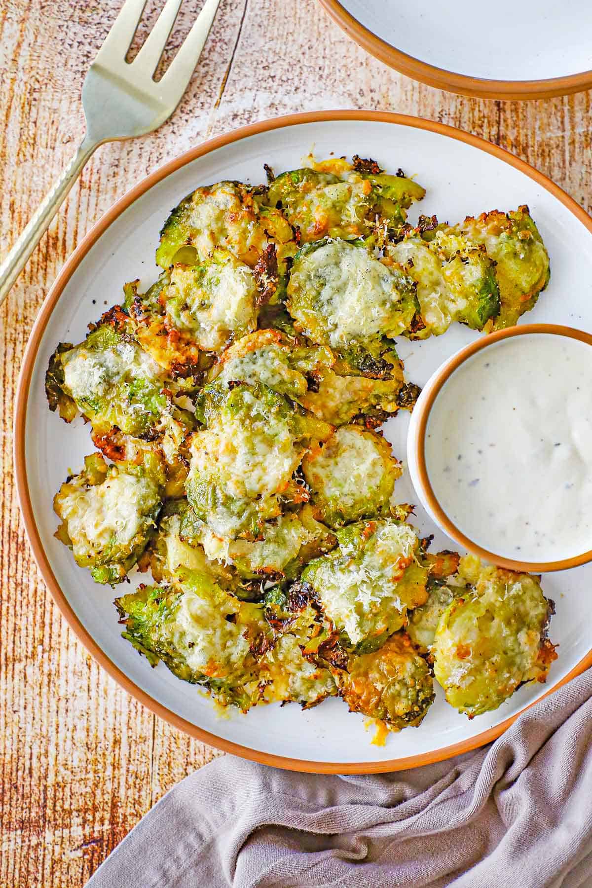An overhead view of a round white plate filled with a serving of Parmesan Smashed Brussels Sprouts in a pile next to a round bowl filled with homemade Ranch dressing.