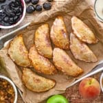 A large baking sheet lined with crumpled brown paper and topped with rows of Southern Fried Fruit Pies surrounded by fruit fillings including cherry, apple, and peach.