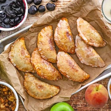 A large baking sheet lined with crumpled brown paper and topped with rows of Southern Fried Fruit Pies surrounded by fruit fillings including cherry, apple, and peach.