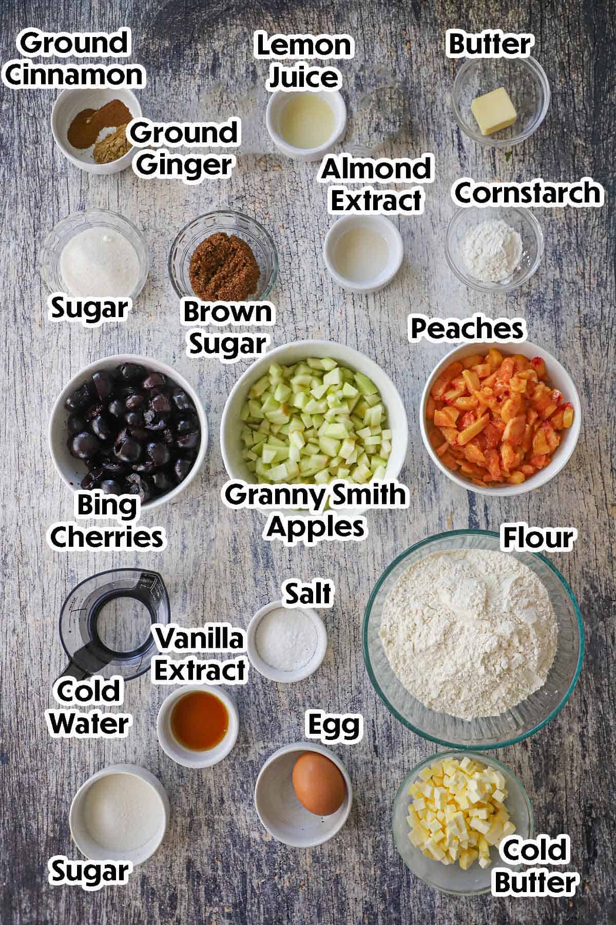 An arrangement of ingredients for Southern Fried Fruit Pies on a grey wooden background including peaches, apples, cherries, flour, butter, an egg, sugar, cornstarch, lemon juice, water, and spices.