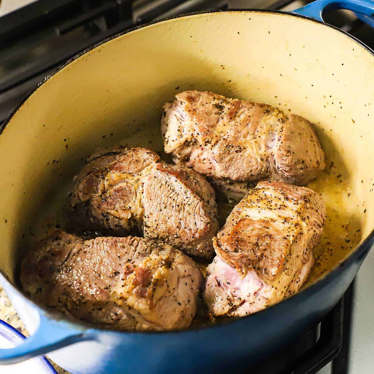 Four pieces of seasoned pork shoulder being seared in oil in a large oval Dutch oven on a gas stove.