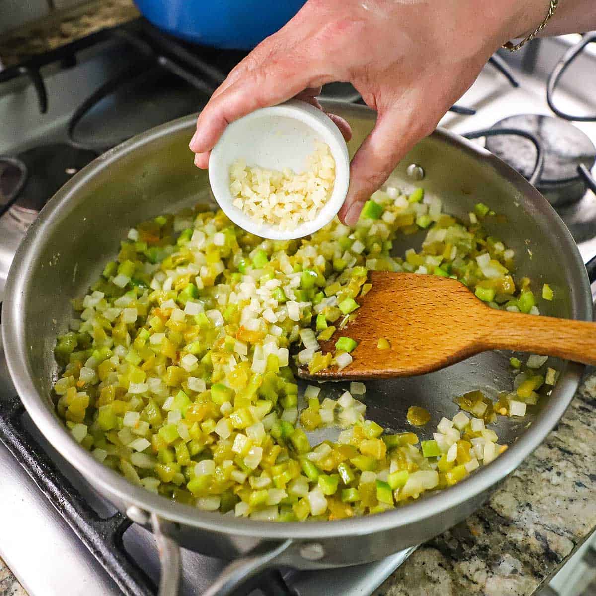 A person transferring minced garlic from a small bowl into a large skillet filled with sautéed onions and green bell peppers over a gas stove.