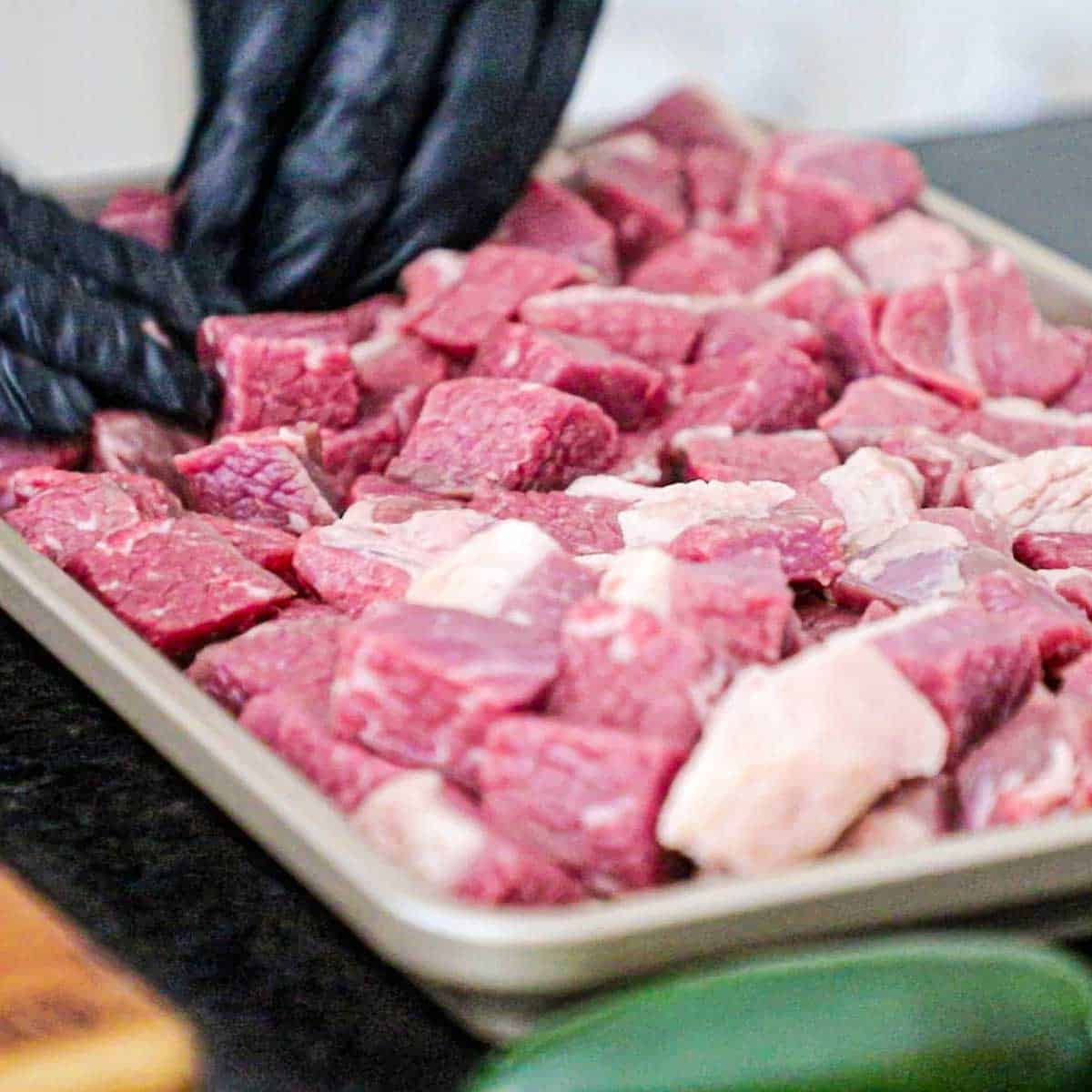 A person placing cubed pieces of beef brisket and pork shoulder onto a rimmed baking sheet.