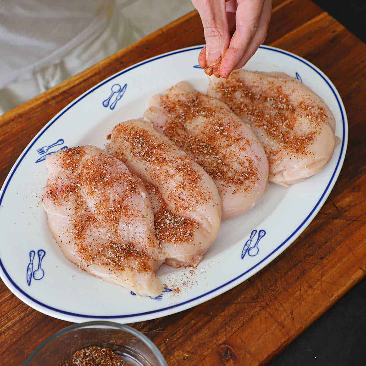 A person using his fingers to sprinkle taco seasoning all over raw skinless chicken breasts on an oval platter on a wooden cutting board.