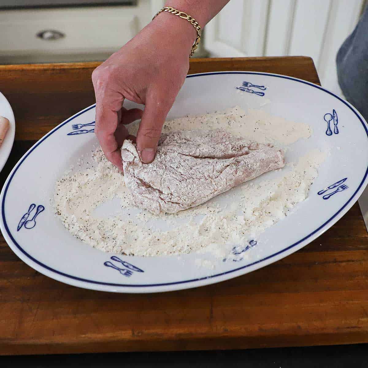 A person dredging a thin boneless chicken cutlet through seasoned flour on an oval platter.