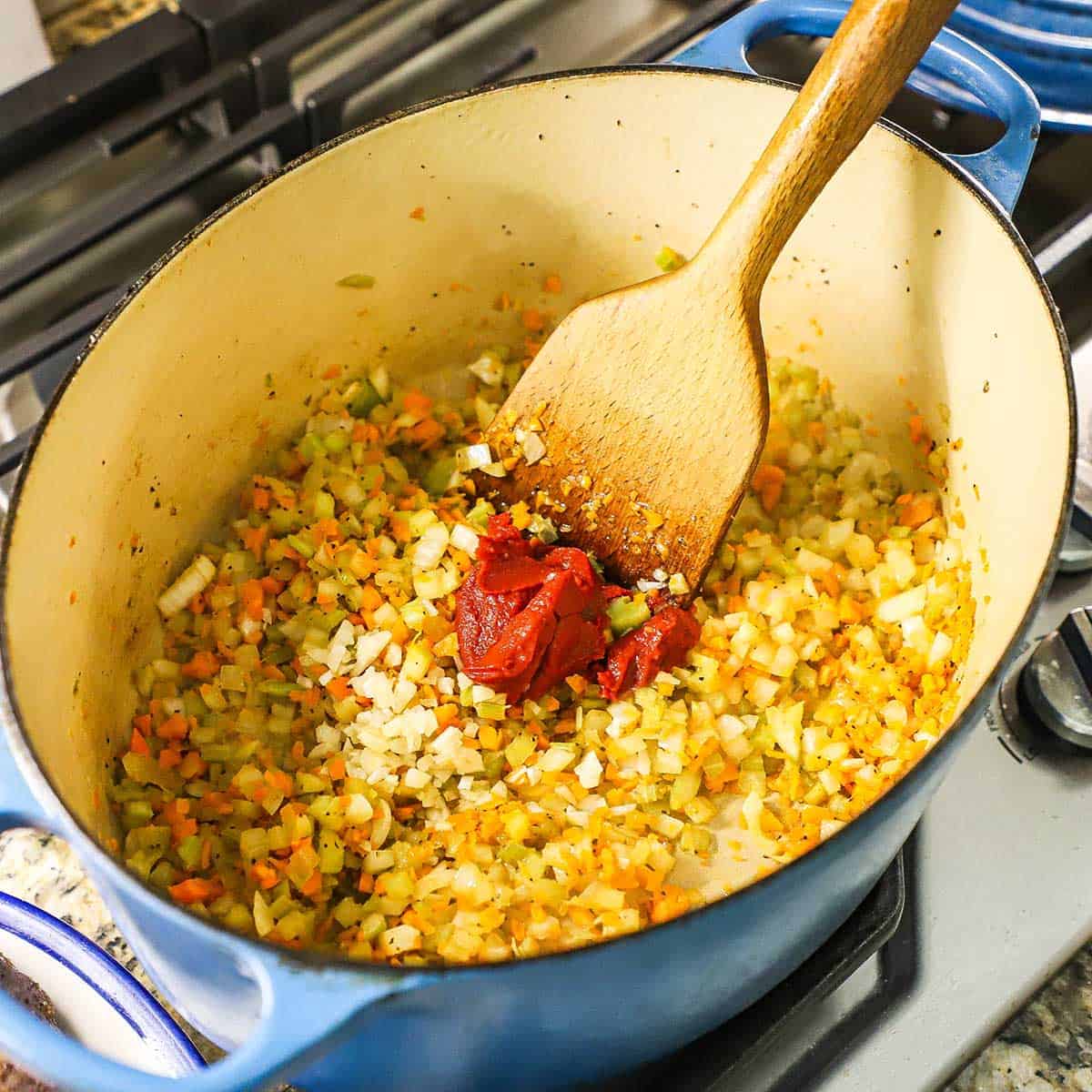 A person using a wooden spatula to stir tomato paste into sautéed chopped vegetables in a large oval Dutch oven on a gas stove.