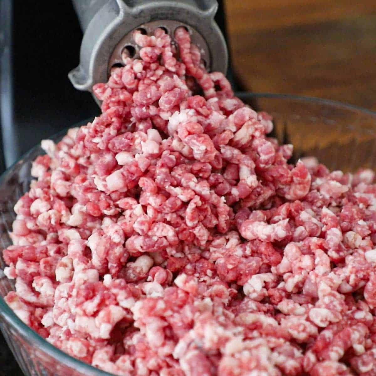 A close-up view of chilled beef brisket and pork shoulder being ground through an electric meat grinder into a large glass bowl.