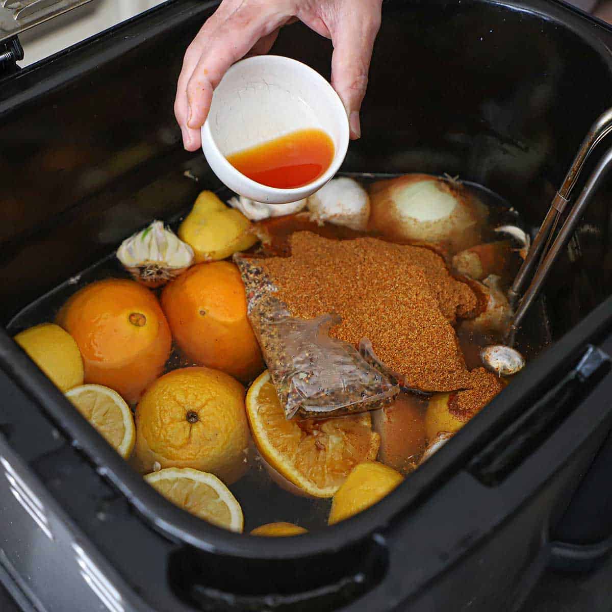 A person pouring a shrimp and crab boil concentrated liquid into a turkey fryer containing halved oranges, lemons, onions, garlic, and a boil-in-a-bag seasoning and a pile of seafood boil spices.