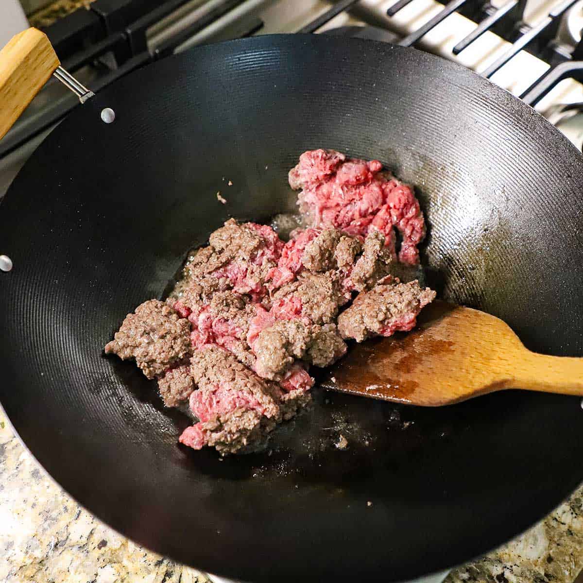 A person using a wooden spatula to flip chunks of partially cooked ground beef in a large wok on a gas stove.