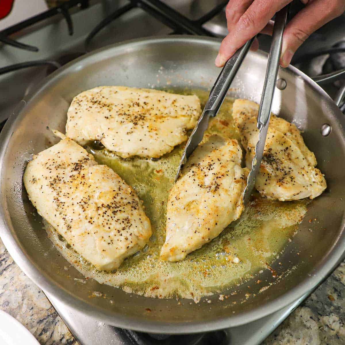 A person searing four floured, seasoned chicken breast in simmering oil and butter in a large stainless steel skillet on a gas stove.