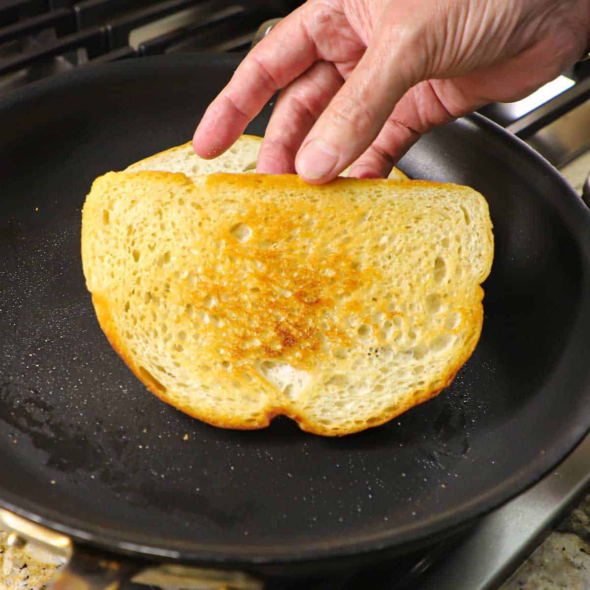 A person flipping a slice of sourdough bread that is being toasted in a large non-stick skillet with butter on a gas stove.