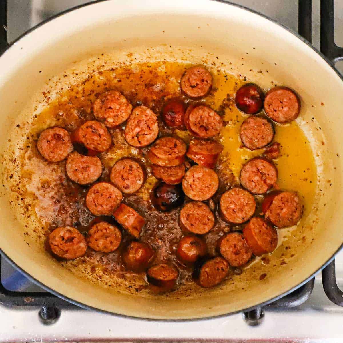 Sliced andouille sausage being seared in oil in a large Dutch oven on a gas stove.