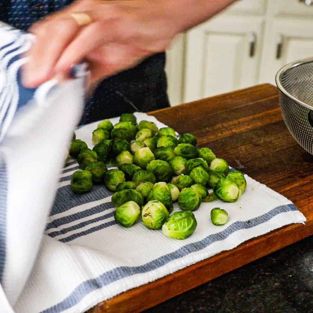 A person using a kitchen towel to dry softened Brussels sprouts on a wooden cutting board.