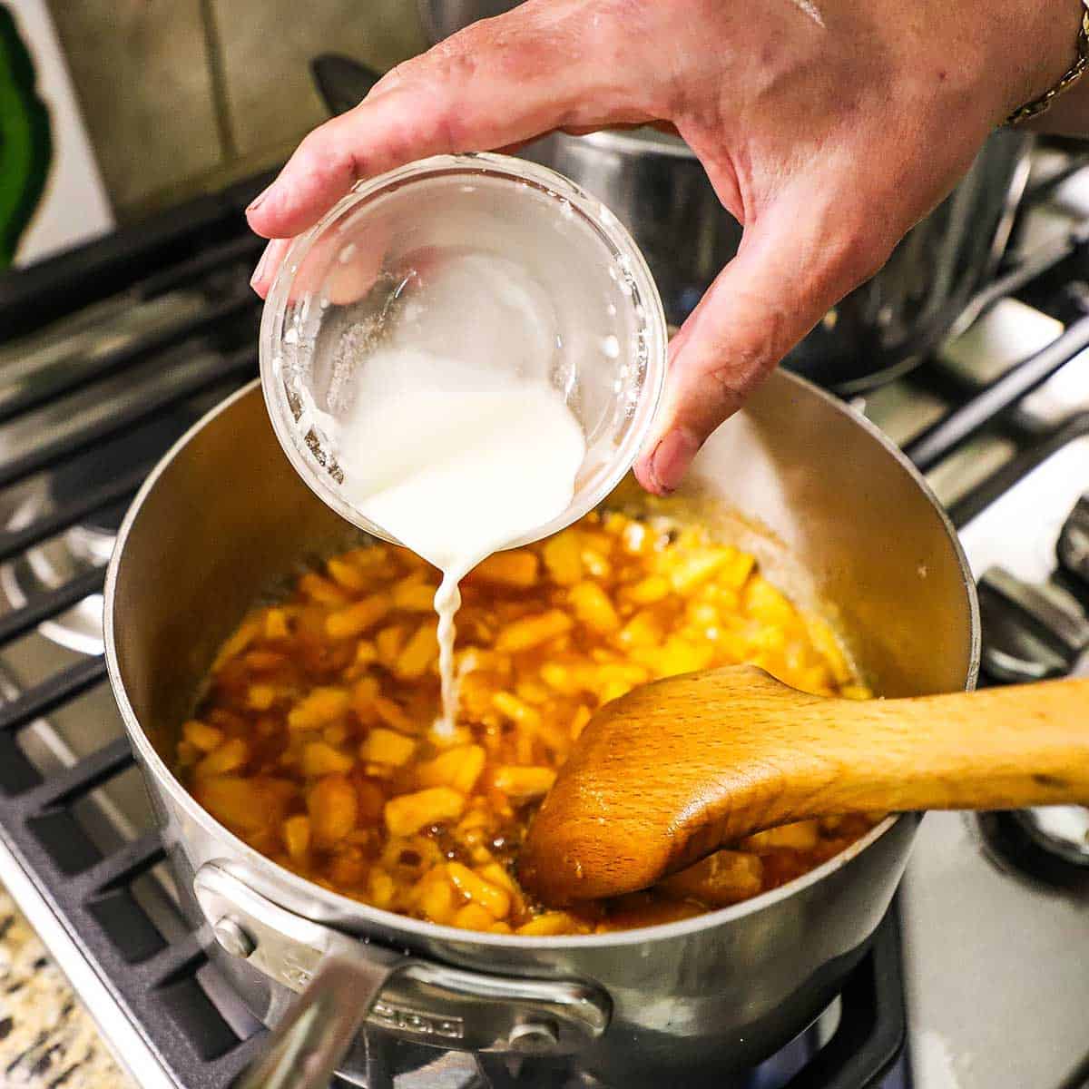 A person pouring a cornstarch slurry from a small glass bowl into a saucepan that is filled with a simmering peach fruit pie filling on a gas stove.