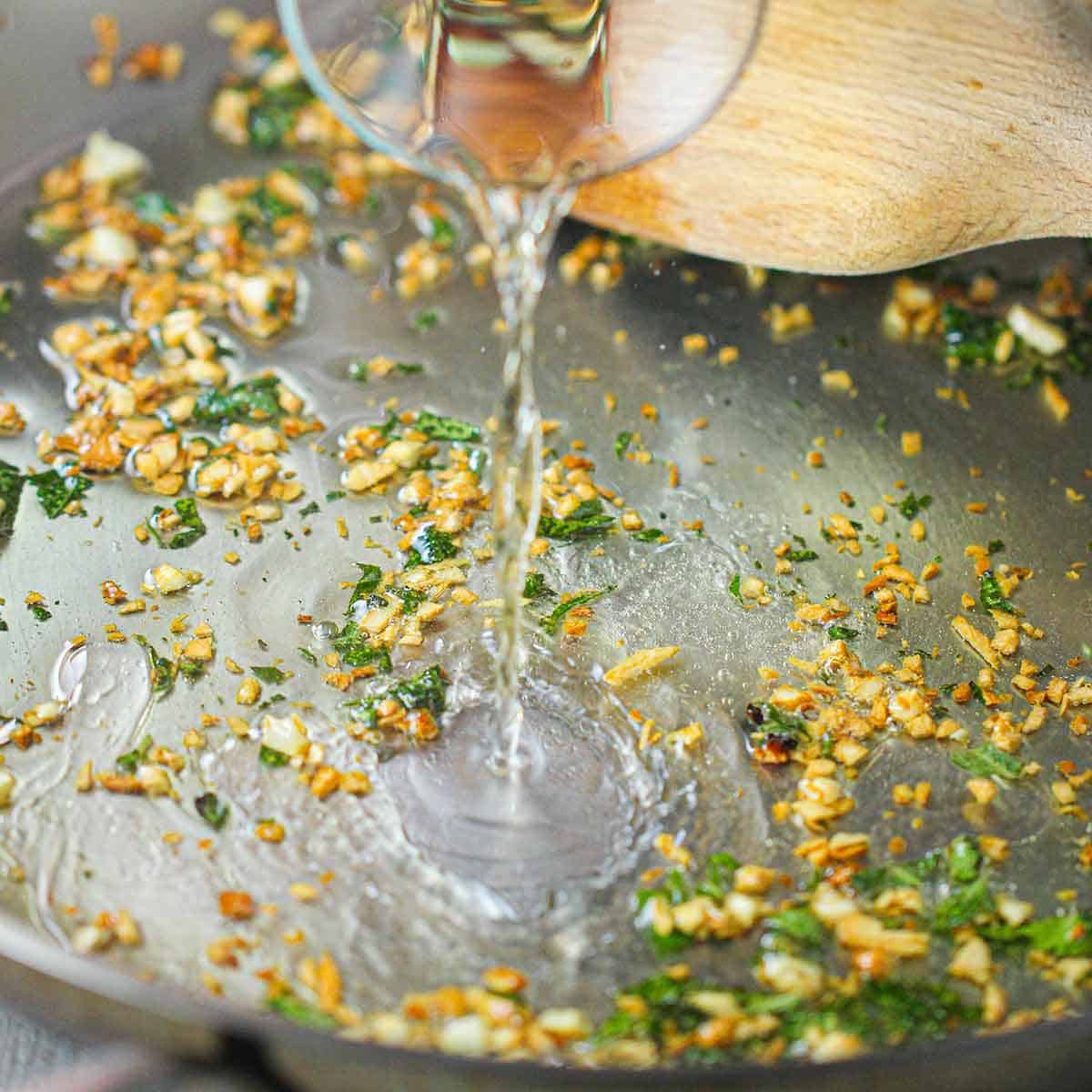 A person pouring white wine from a small carafe into a skillet filled with sautéed garlic and chopped fresh sage.
