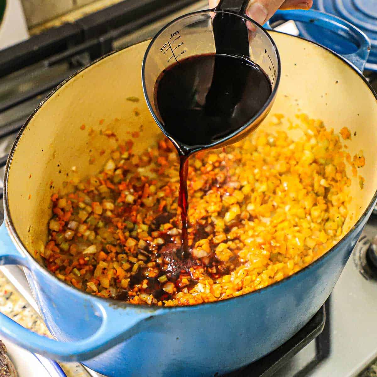 A person pouring red wine from a plastic measuring cup into a large oval Dutch oven that is filled with sautéed chopped vegetables.